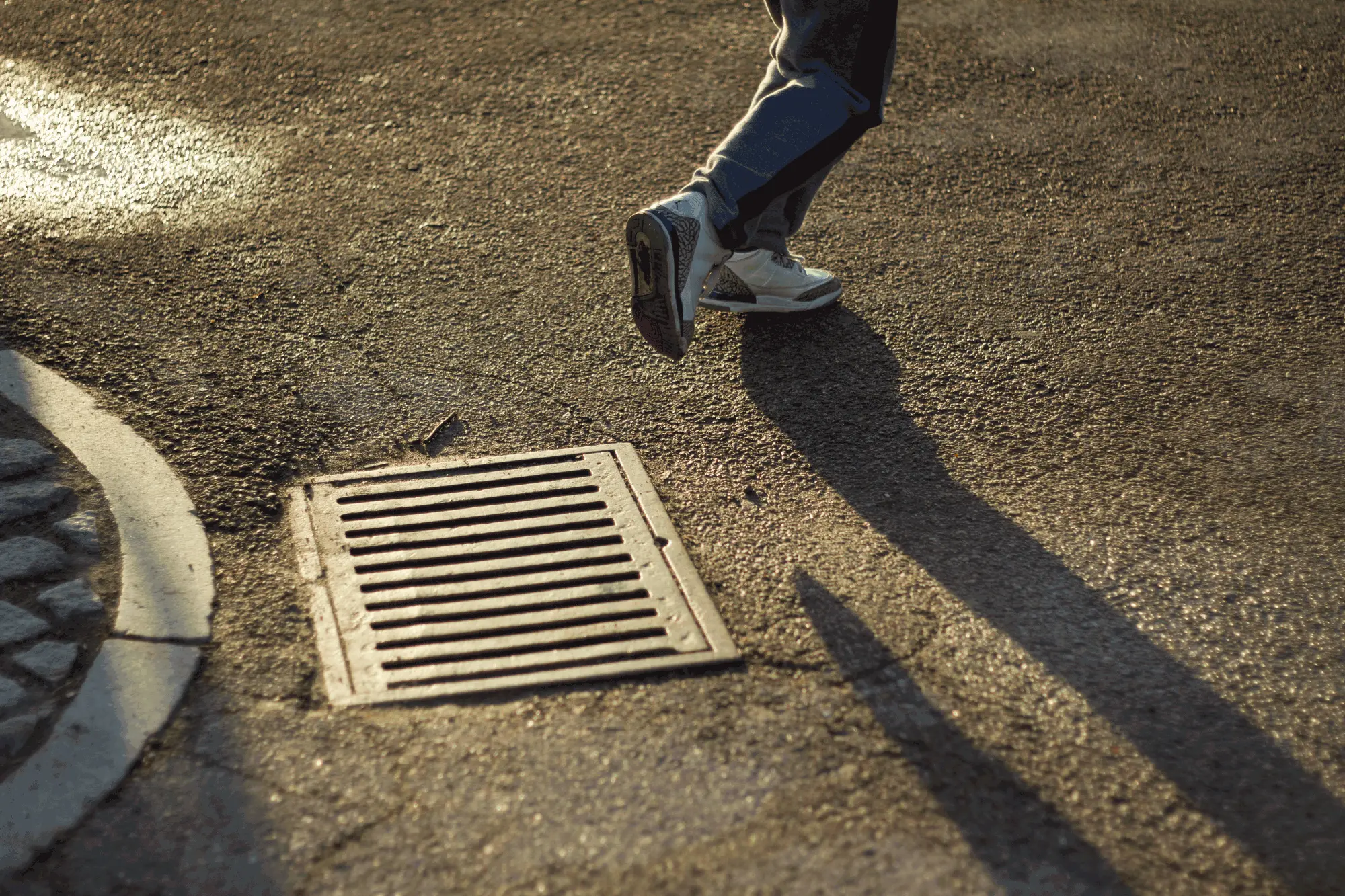 Someone in sneakers stepping past a storm drain, casting long shadows in low evening sun
